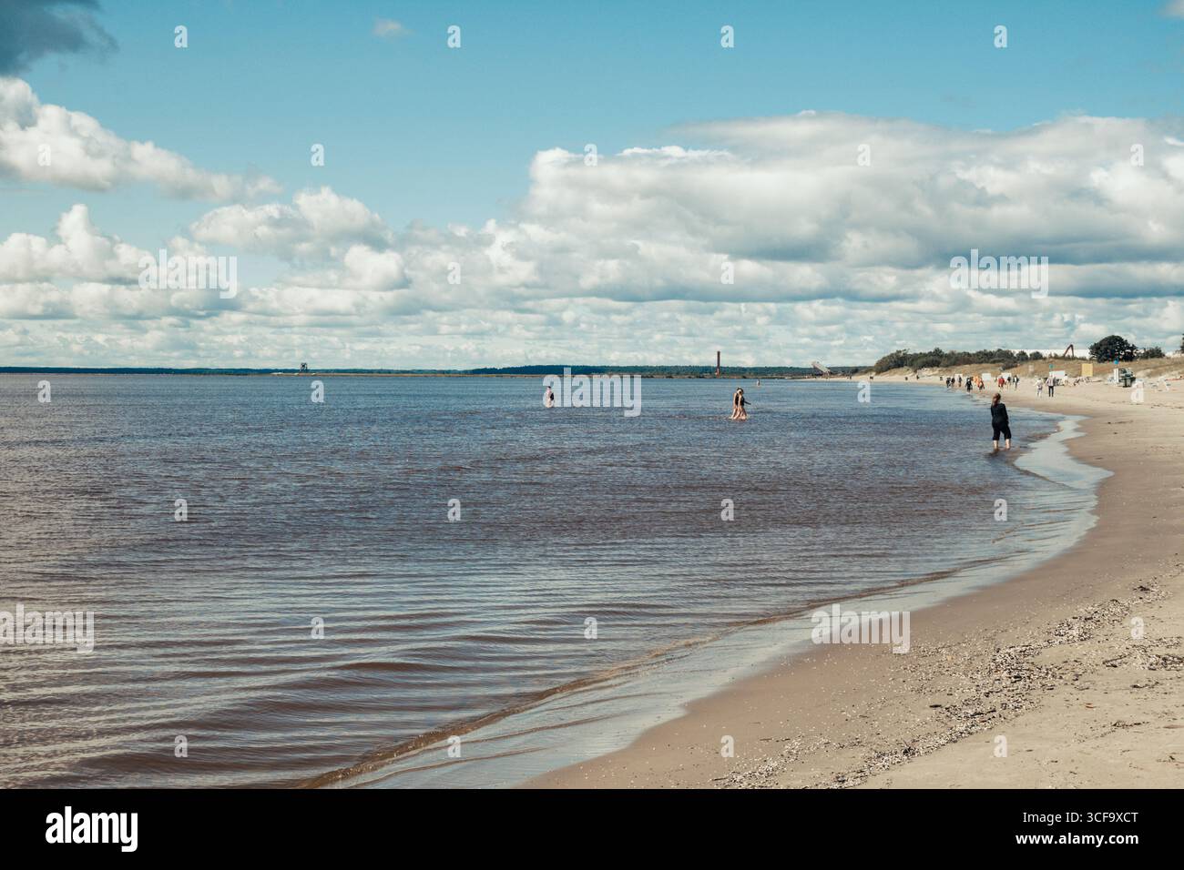 People walking along a sandy beach near the shoreline, with the water gently meeting the shore in a summer day Stock Photo