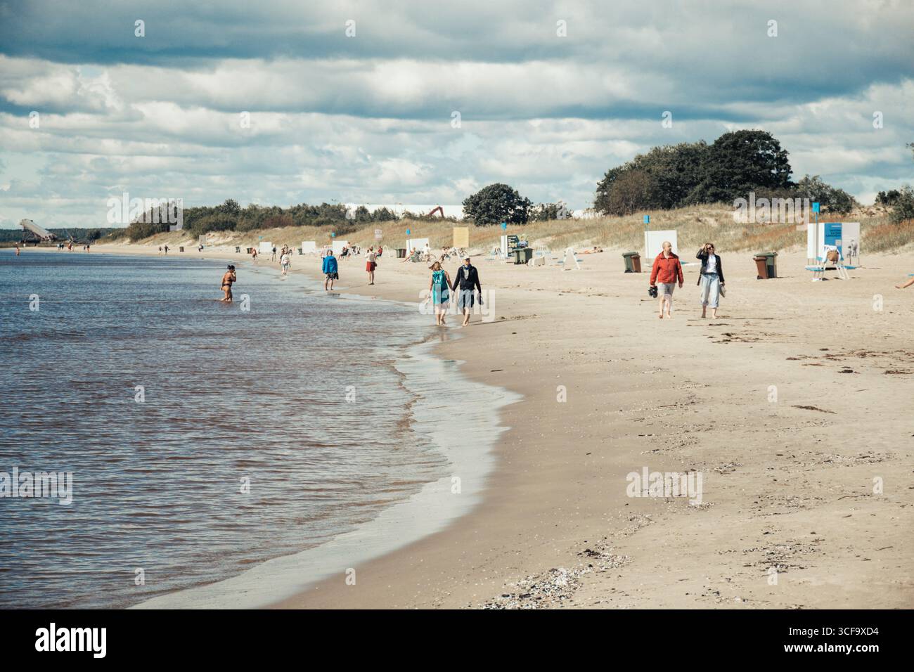 People walking along a sandy beach near the shoreline, with the water gently meeting the shore in a summer day Stock Photo