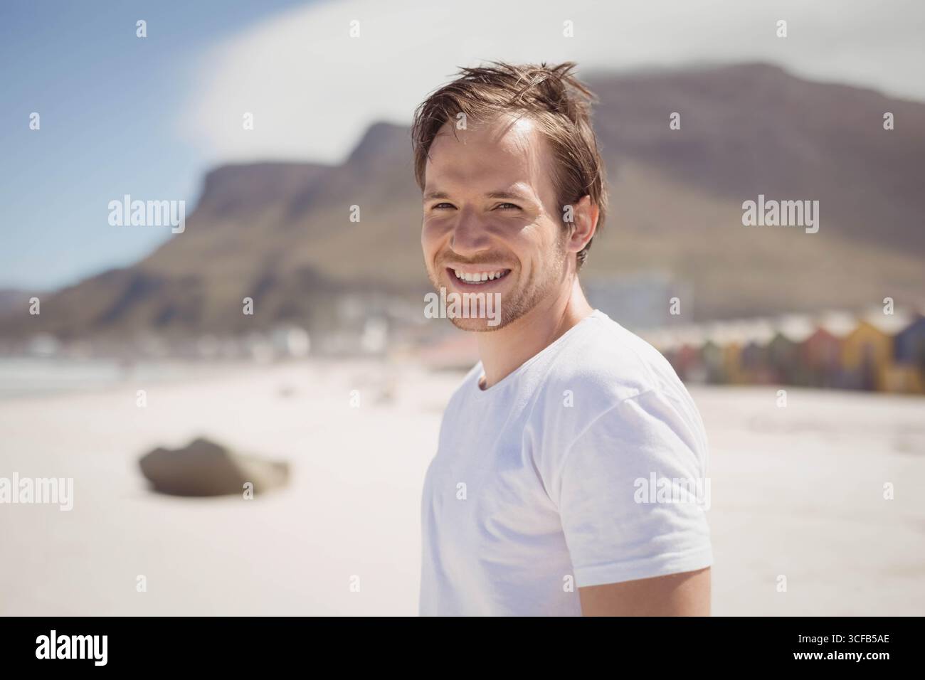 Man standing on sandy beach wearing white t-shirt beside boulder, colorful beach huts, copy space Stock Photo