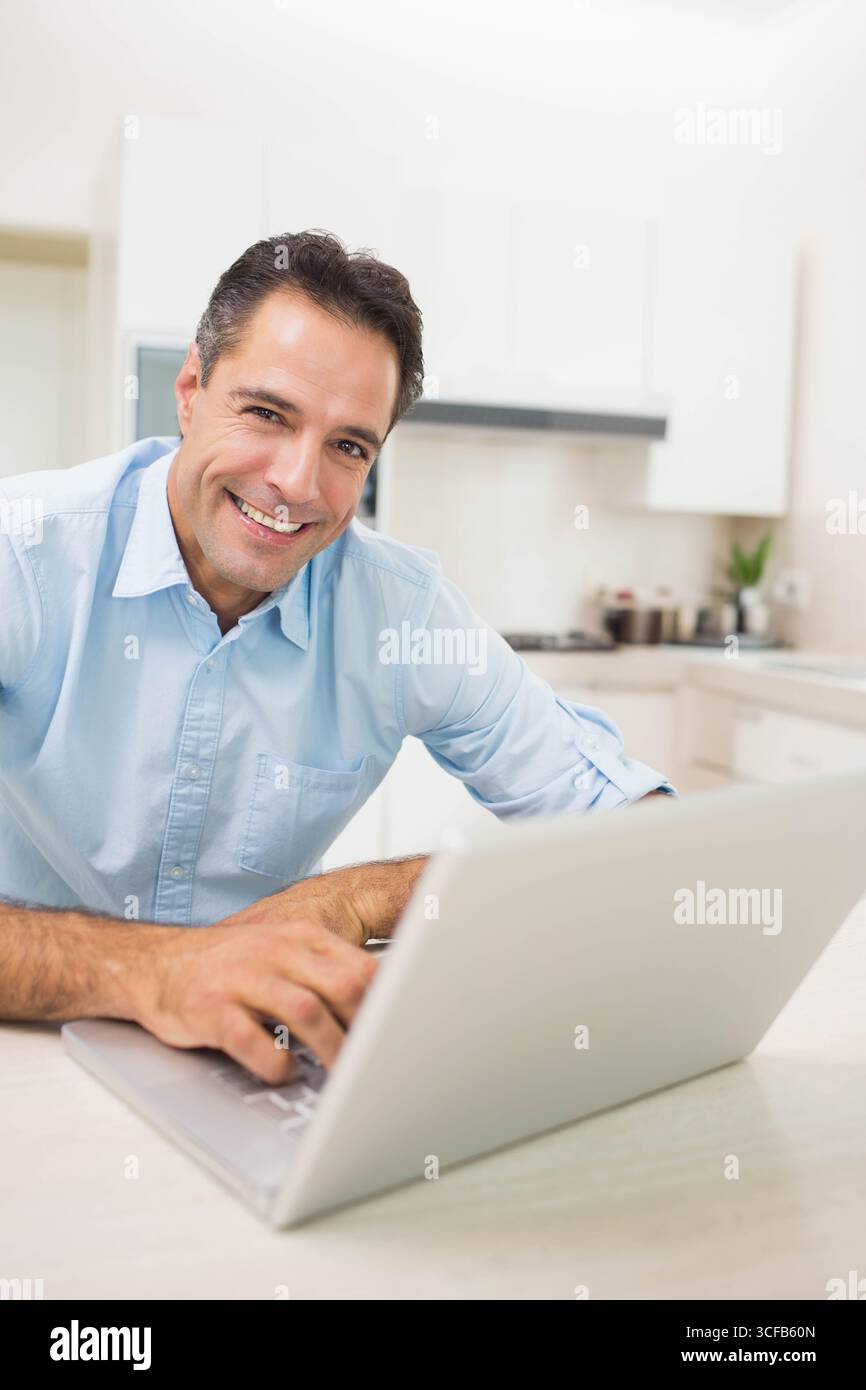 Middle-aged man in casual shirt leaning over kitchen counter using silver laptop next to spice jars Stock Photo