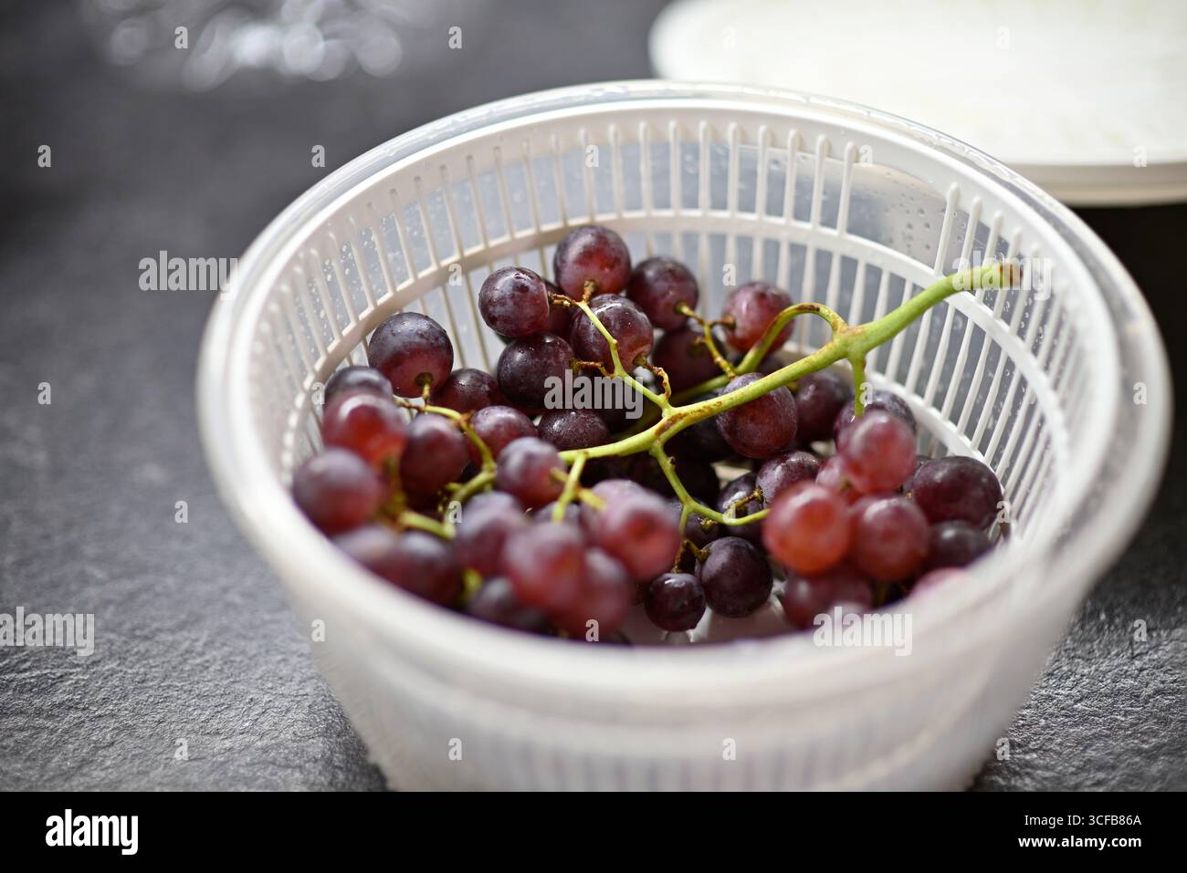 Fresh red grapes in a white plastic colander on a dark kitchen surface. Stock Photo