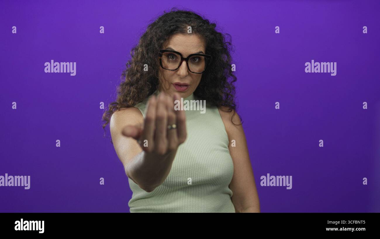Young woman wearing glasses and green top beckons hand forward in purple studio; friendly invitation. Stock Photo
