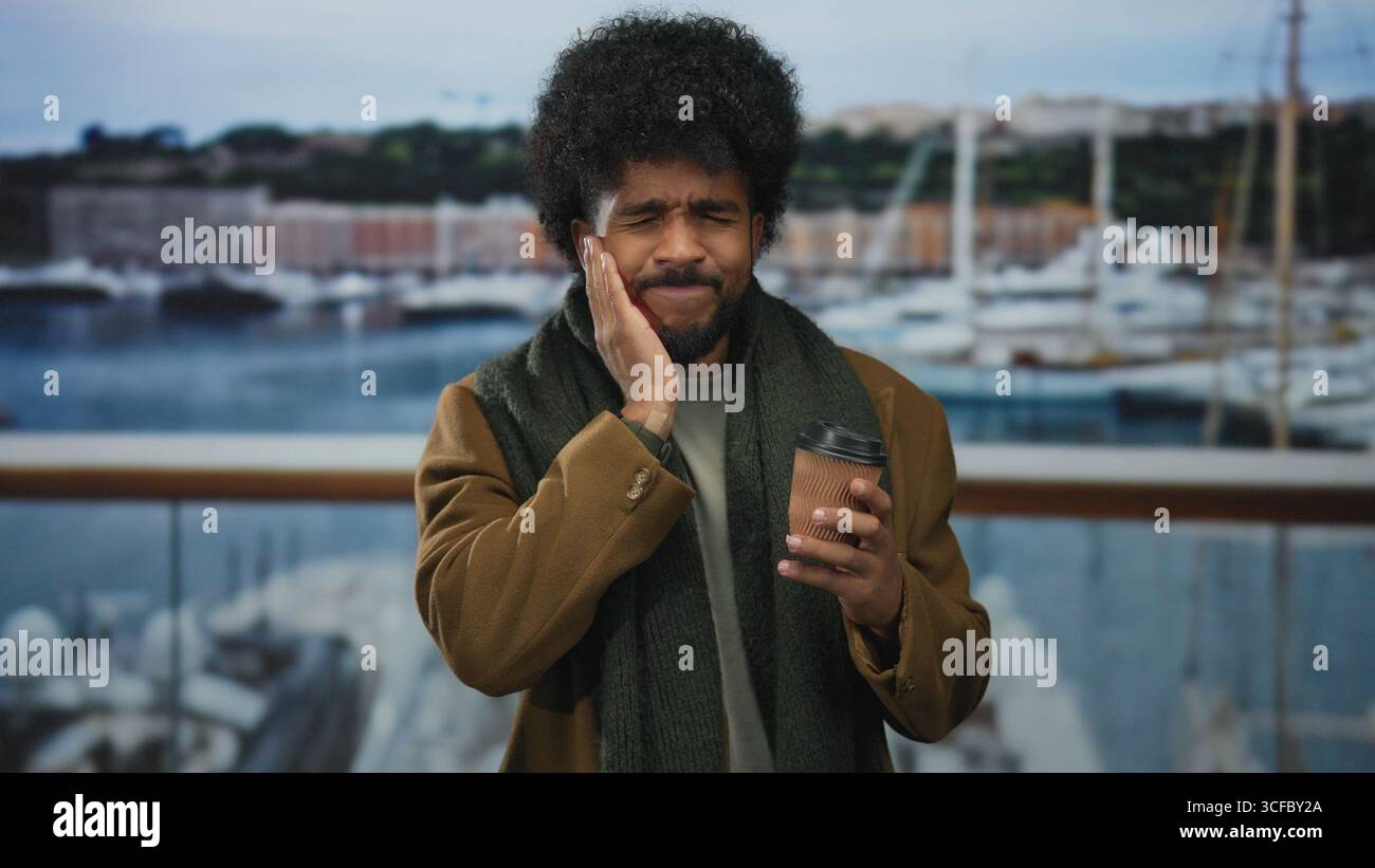 Man stands outdoors by the seaside holding coffee near boats, expressing tooth pain while touching jaw against a backdrop of a port and calm sea. Stock Photo