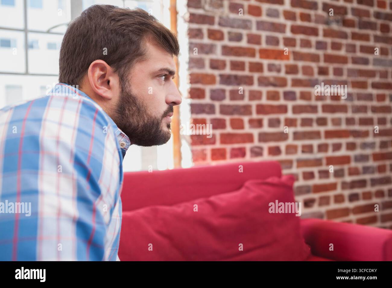 Man sitting on red sofa beside window in lounge with brick wall, gazing right, copy space Stock Photo