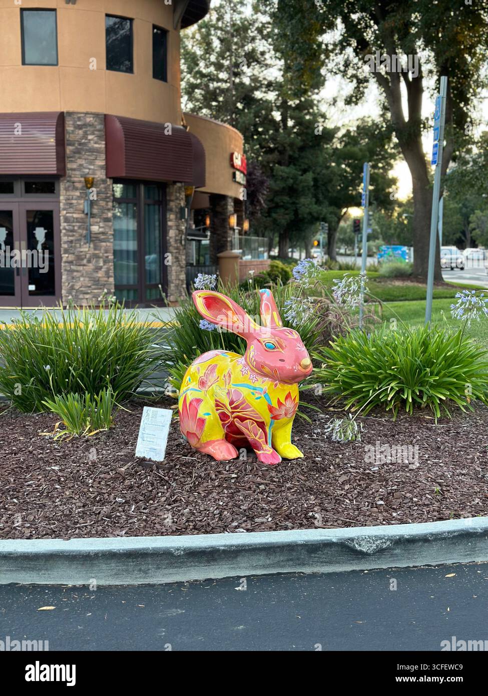 A brightly painted rabbit sculpture is displayed as a public art installation in a landscaped street median near shops - Smartphone Captured Stock Image
