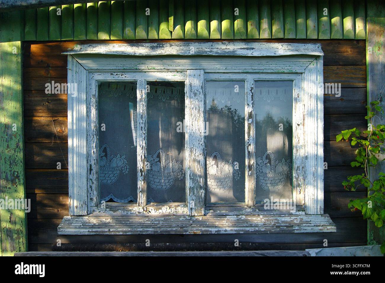 A wooden house in Russia in the summer, lace curtains on the window, the paint peeled off, sun glare on the glass. Comfort, home, village life. Peaceful life - Stock Image
