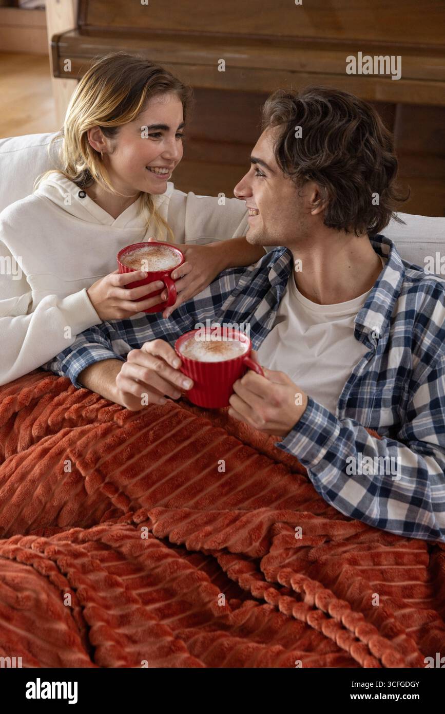 Couple sitting on sofa under red blanket holding coffee mugs in living room by wooden piano Stock Photo
