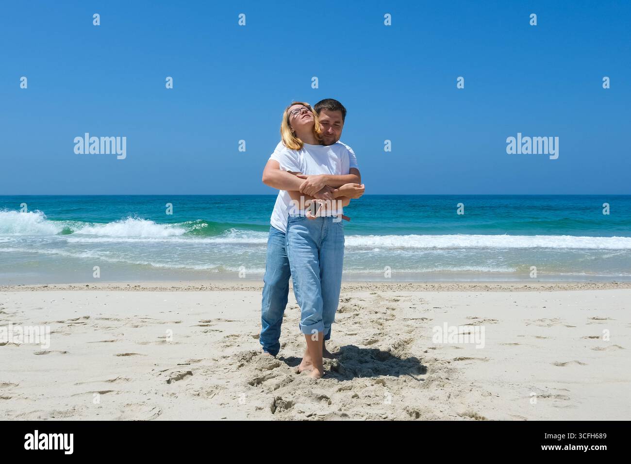 Couple standing barefoot on sandy beach with man hugging woman from ...