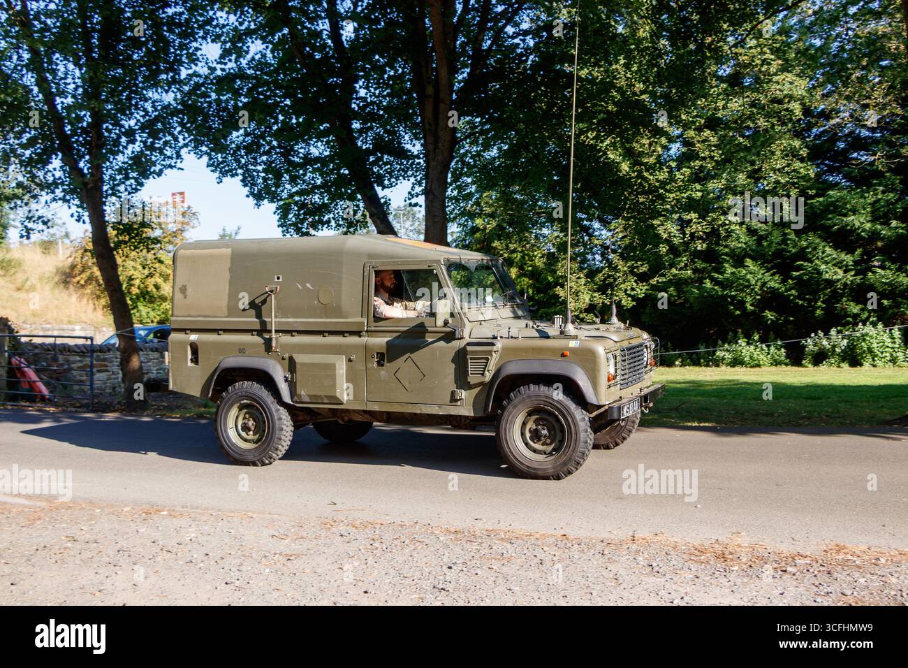 Vintage british army land rover defender military vehicle driving on a rural road surrounded by trees and greenery on a sunny day Stock Photo