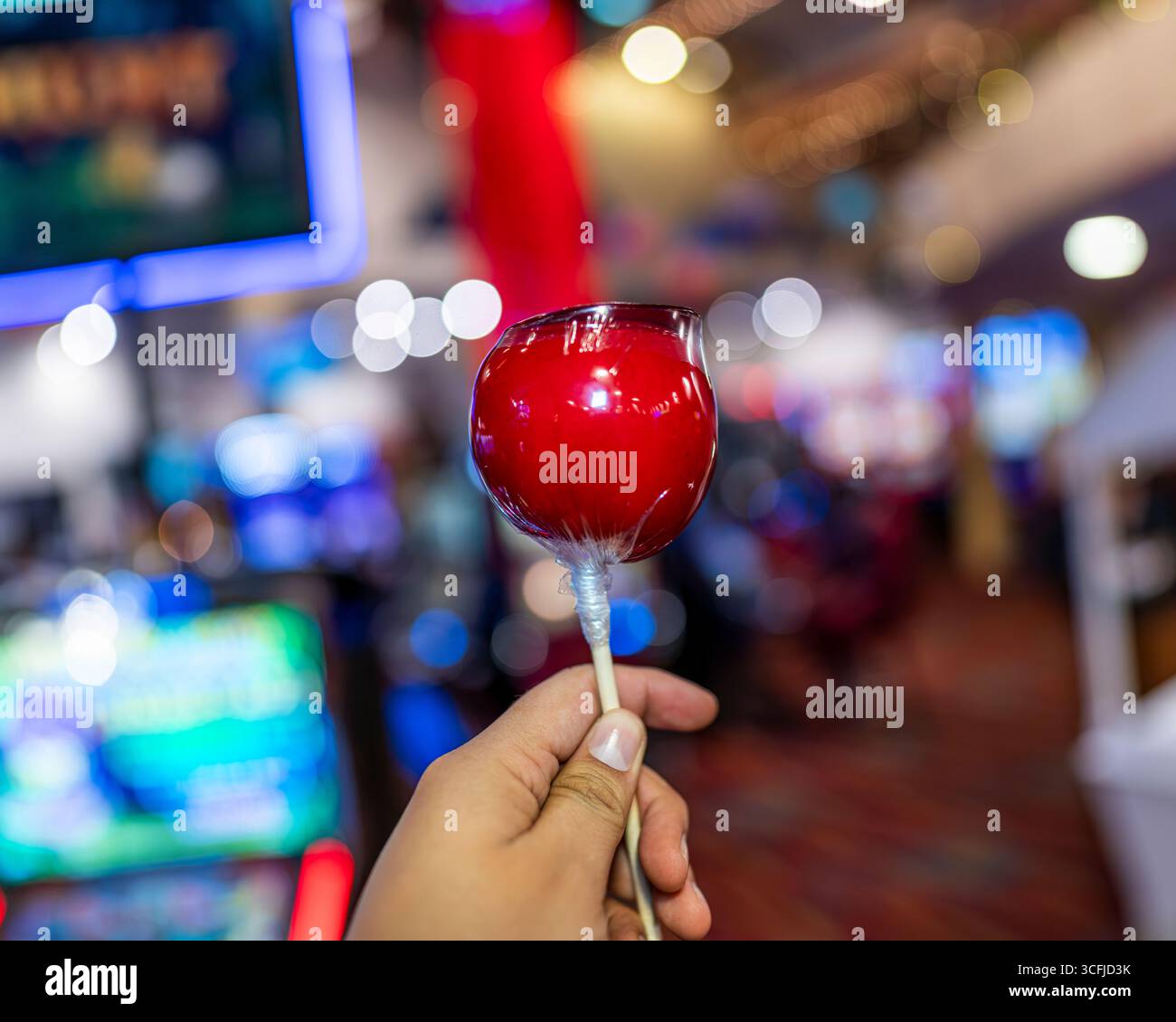 Hand holding a red candy apple on stick inside casino with colorful blurred lights in background, symbolizing dessert, snack, and festive celebration. Stock Photo