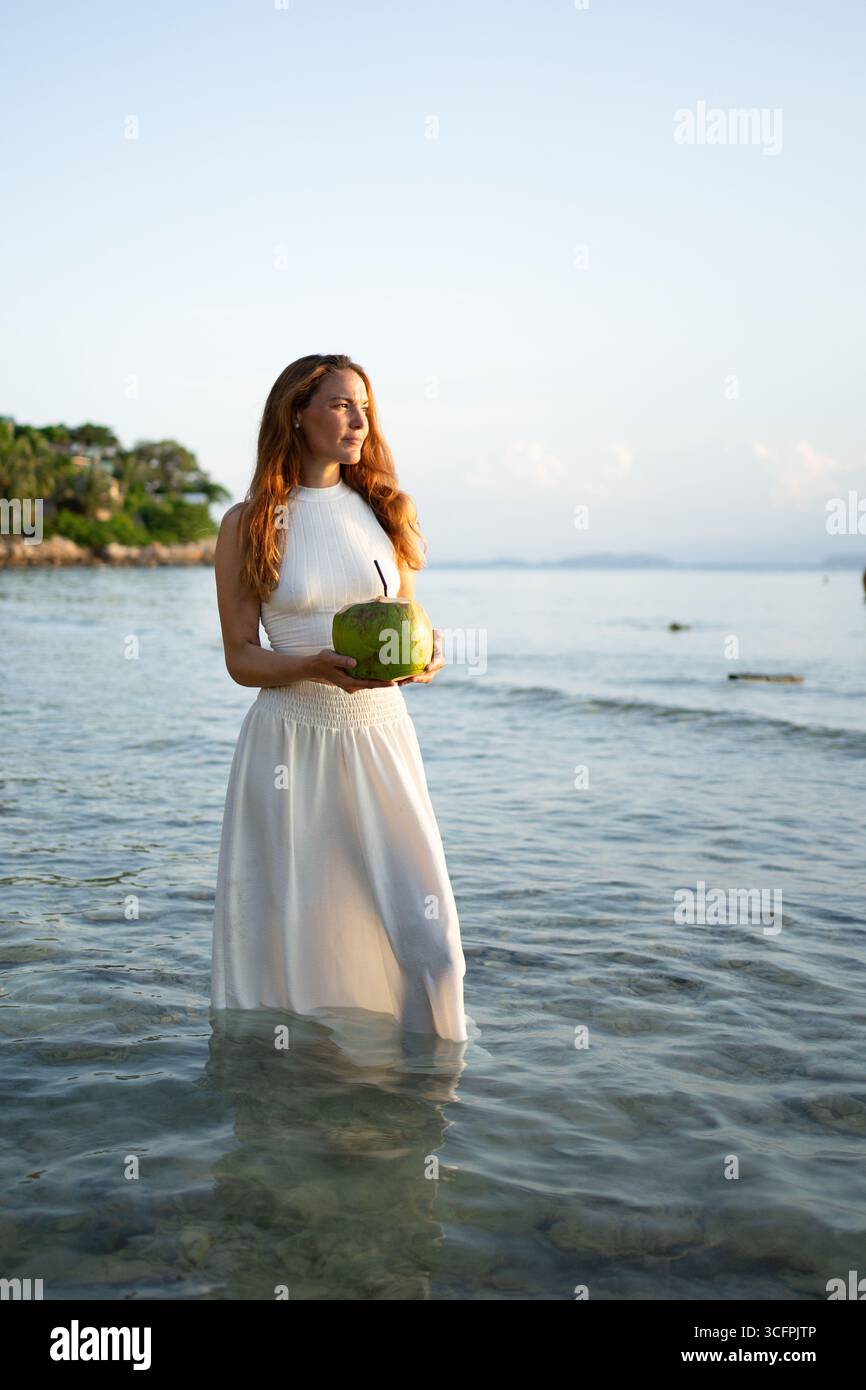 Tourist enjoying refreshing coconut water while standing in shallow water on a tropical beach at sunset Stock Photo