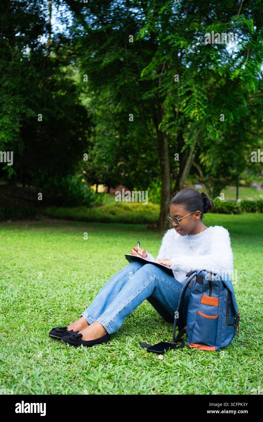 Focused student taking notes in a notebook while sitting on the grass in a park, enjoying the fresh air and serene environment Stock Photo