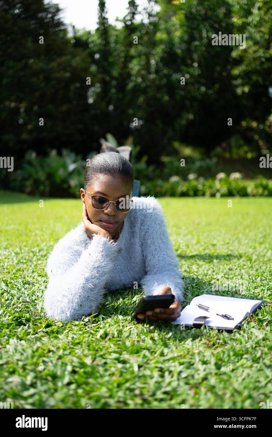 Young african american woman lying on grass using her phone, with a notebook and pen next to her, enjoying studying outdoors Stock Photo