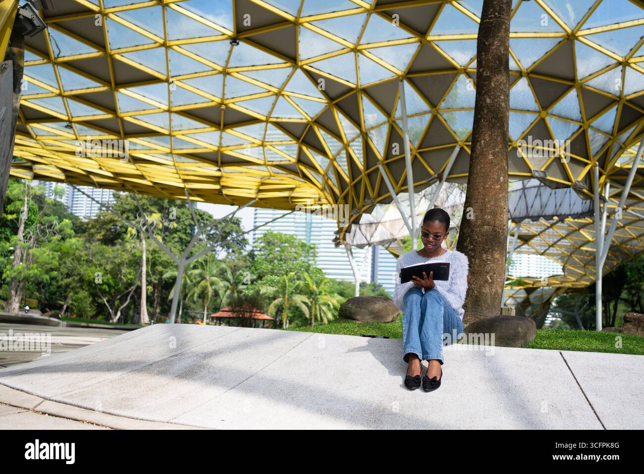 Young woman enjoying reading a book in a peaceful park setting, under a striking yellow canopy structure Stock Photo