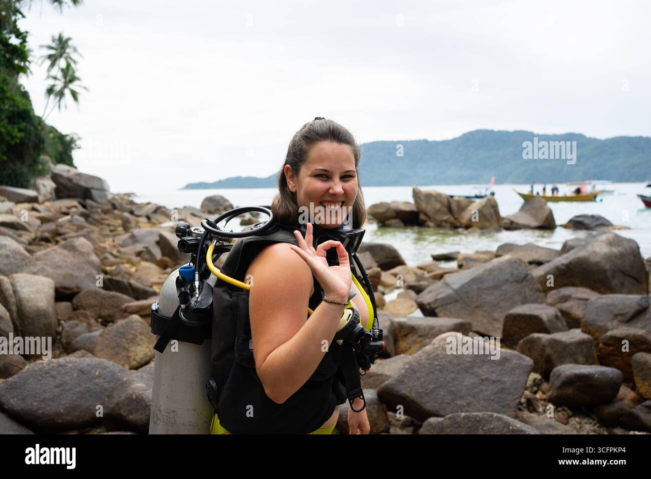 Female scuba diver smiling and gesturing ok sign on a rocky beach, ready for diving Stock Photo