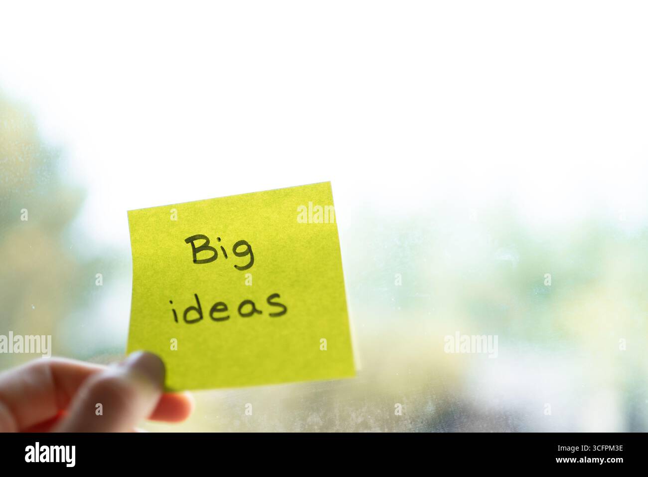Sticky note with big ideas writing held by a hand against blurry window background Stock Photo