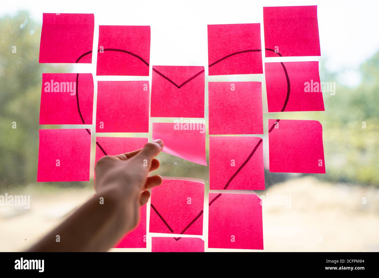 Hand placing pink sticky notes with a heart shape drawn on them onto a glass window, creating a romantic message Stock Photo