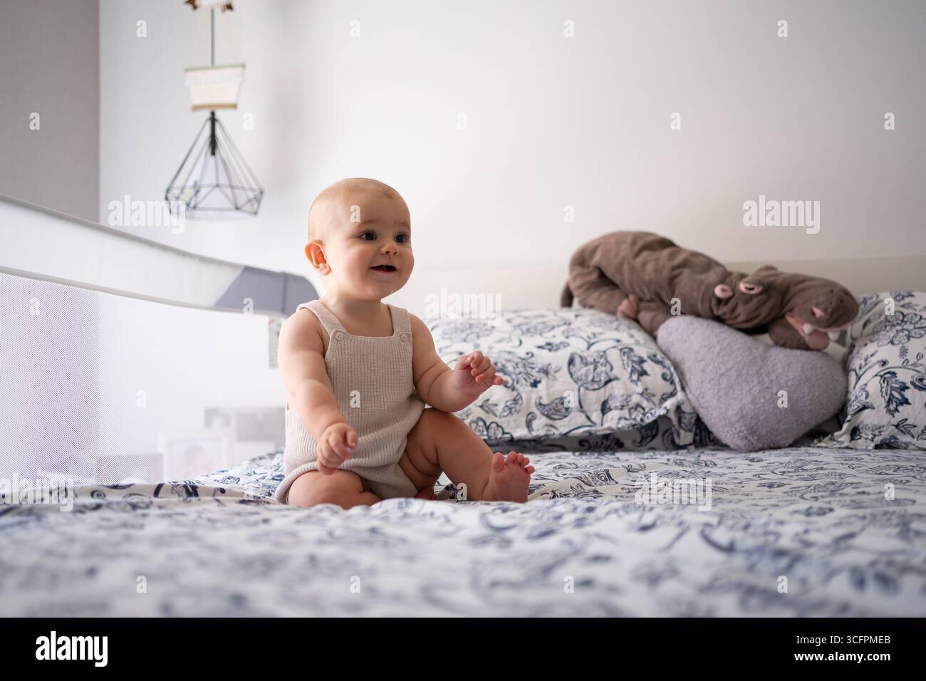 Smiling baby girl sitting on bed playing with plush hippo toy in bedroom Stock Photo