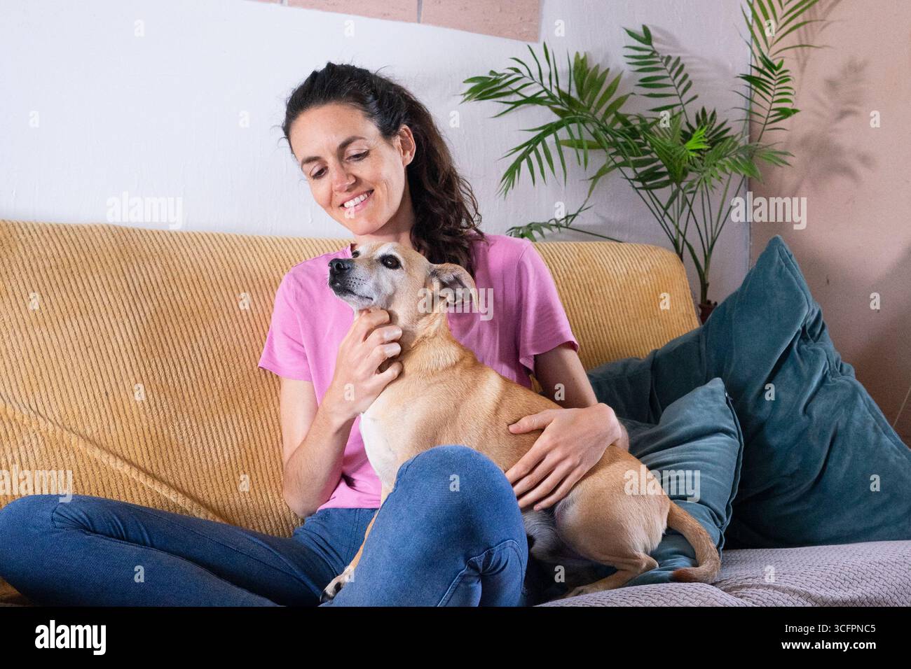 Happy woman is cuddling her small brown dog while sitting on a comfortable sofa in her living room Stock Photo