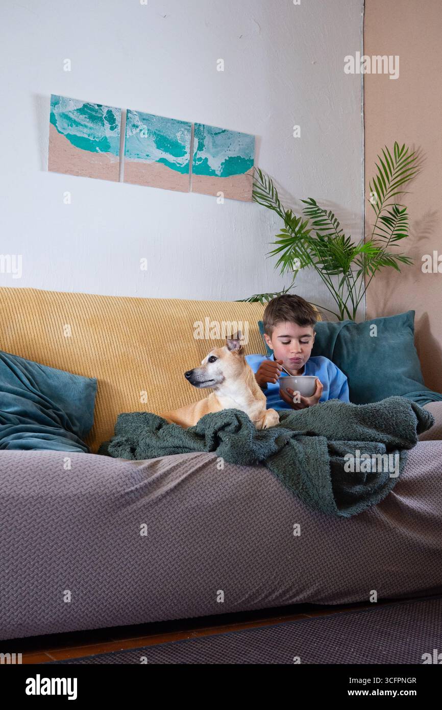 Child eating a bowl of cereals while sitting on a comfortable sofa with his dog Stock Photo