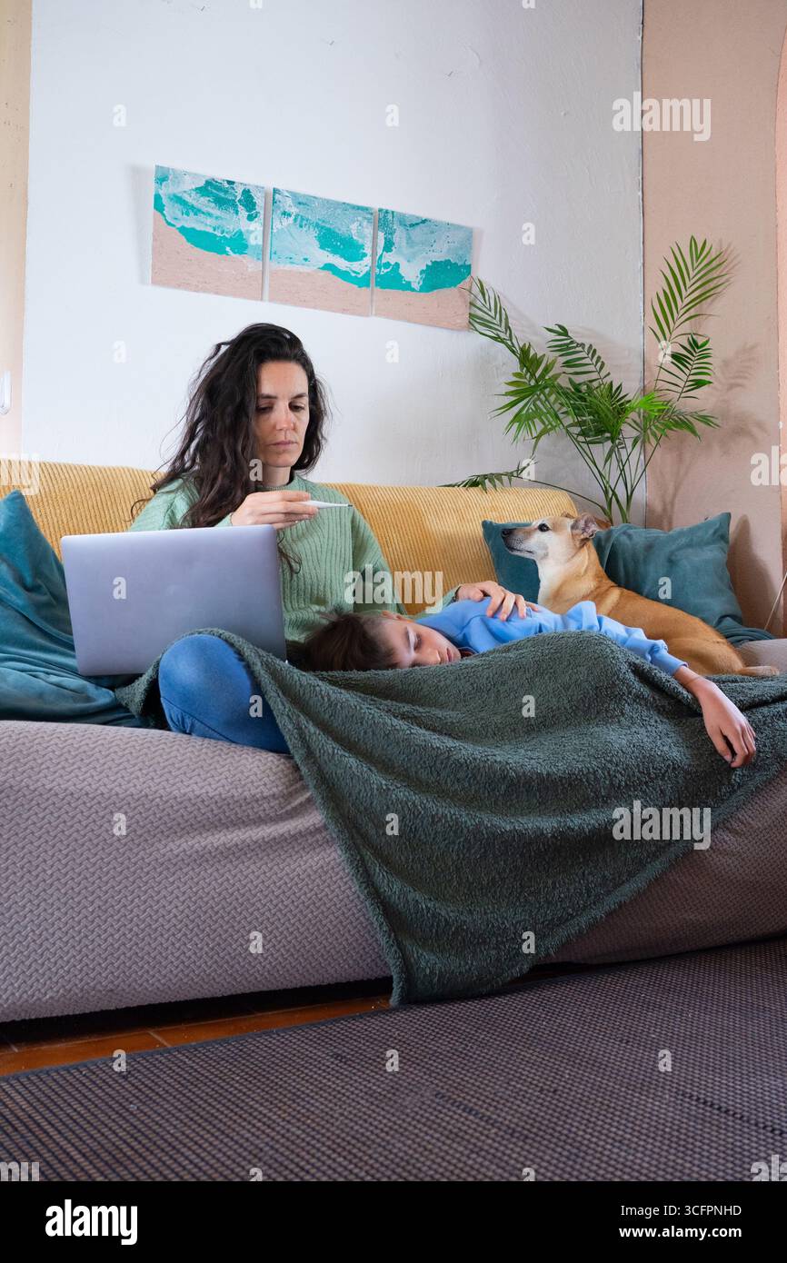 Mother working from home checking temperature of sick child lying on sofa with dog Stock Photo