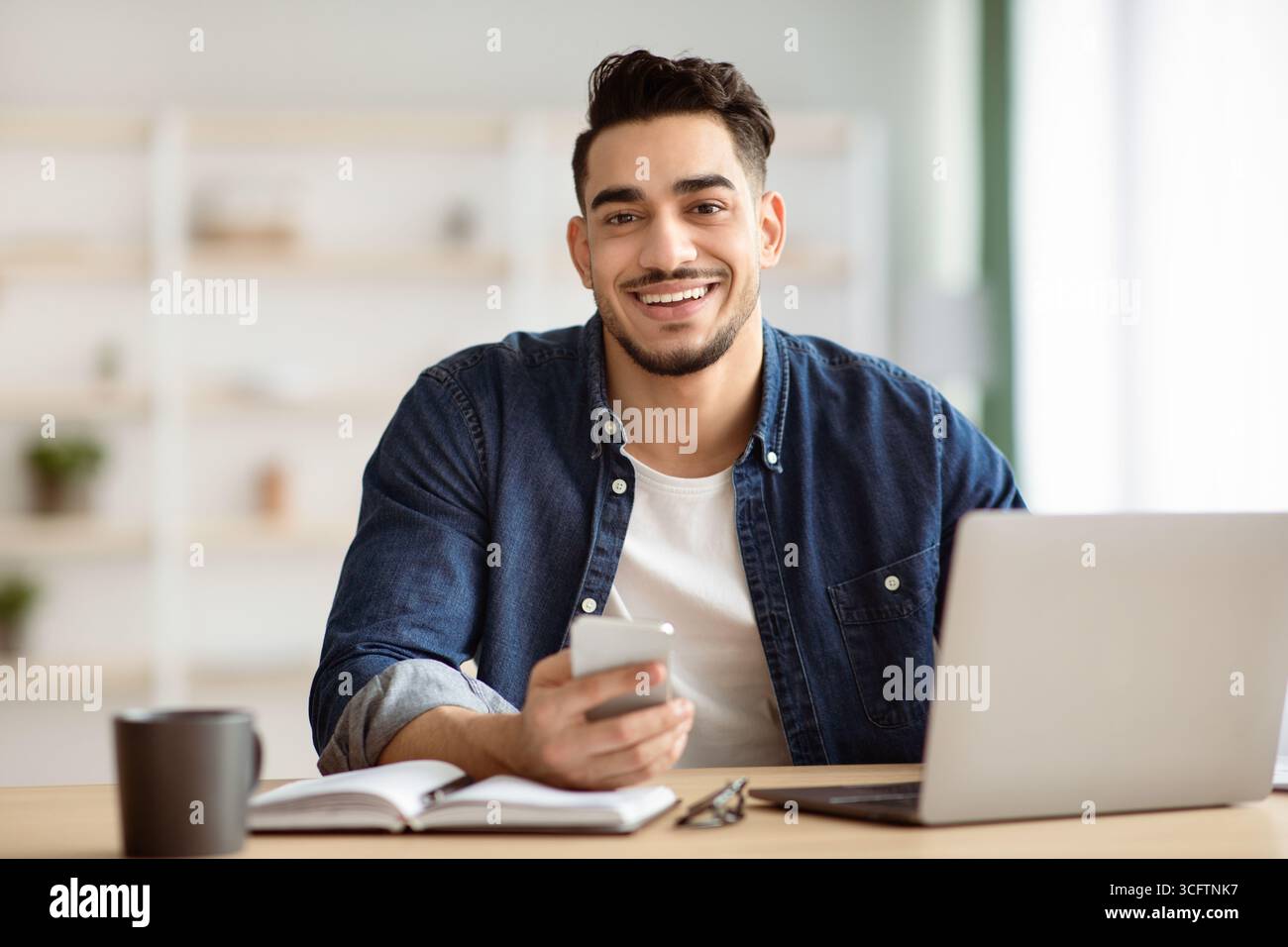 Smiling arab guy using smartphone while sitting at workdesk Stock Photo