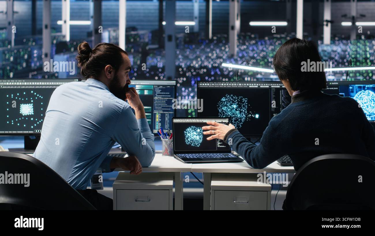 Server room system administrators using AI to review infrastructure diagnostics. IT professionals in data center analyzing performance data using artificial intelligence technology, camera A Stock Photo