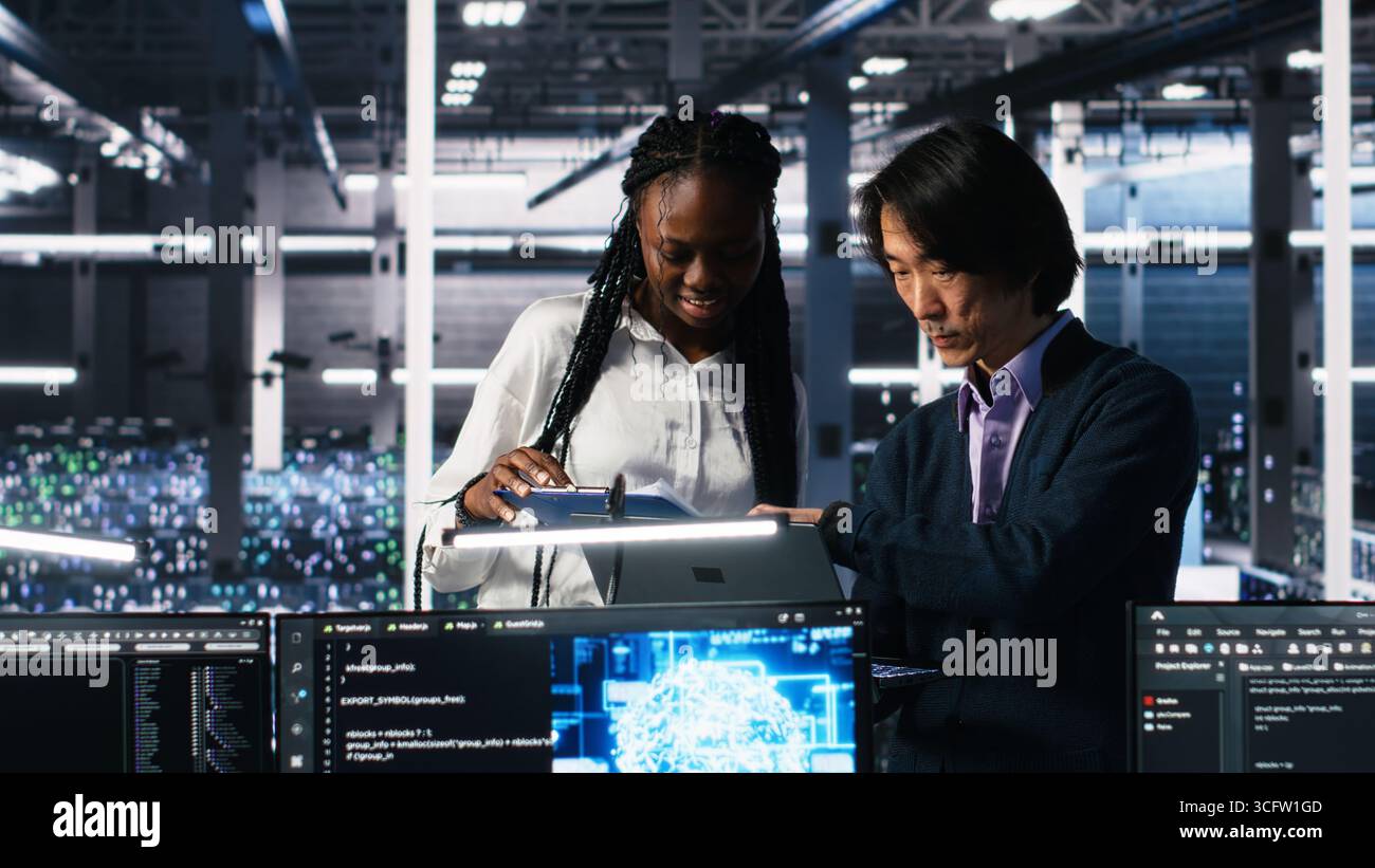 Team of data center engineers looking over paperwork and using deep learning algorithms to analyze datasets. Employees in multiethnic server farm using AI to recognize data patterns, camera A Stock Photo