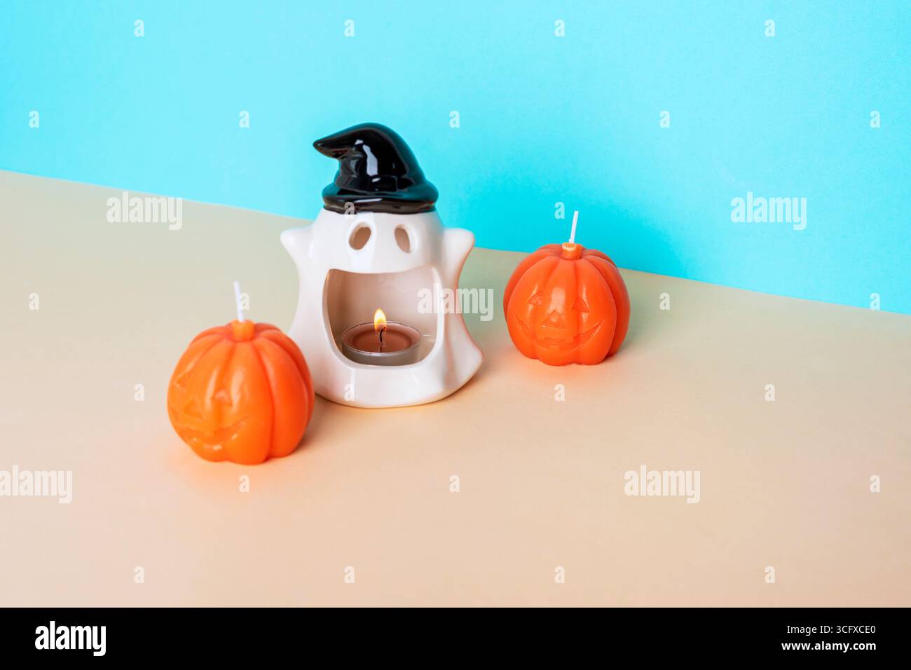 Ceramic ghost candle holder with burning tealight and two pumpkin shaped candles on blue and beige background creating fun Halloween decoration scene. Stock Photo