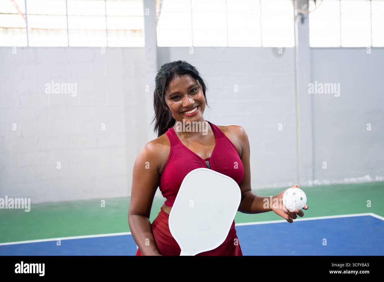 Happy pickleball player holding paddle and ball smiling on indoor court Stock Photo