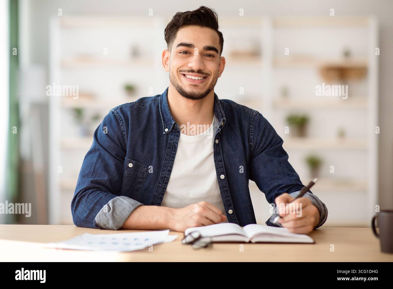 Cheerful arab guy in casual sitting at workdesk, taking notes Stock Photo