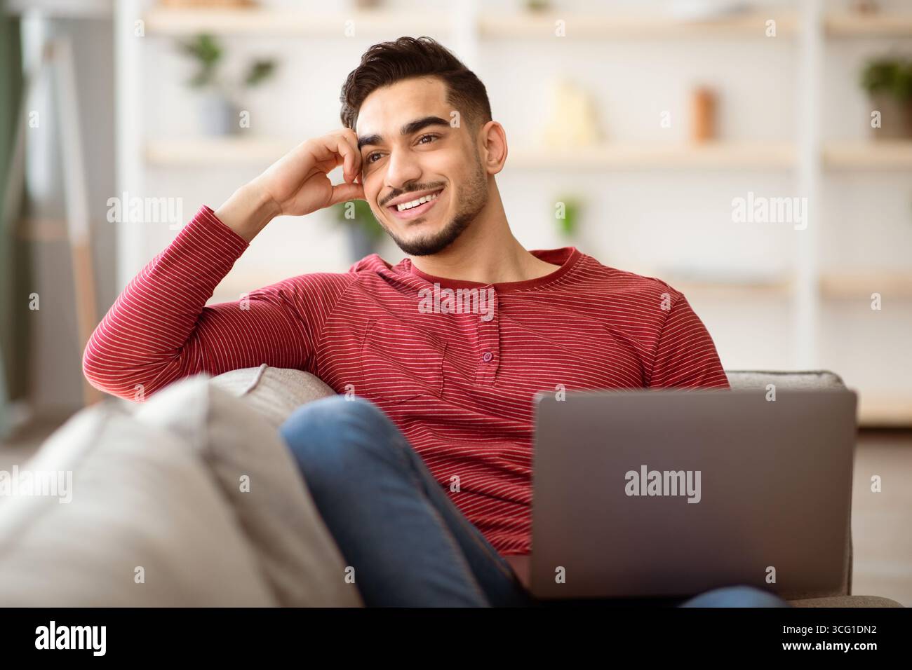 Dreamy arab guy resting on couch at home, using laptop Stock Photo