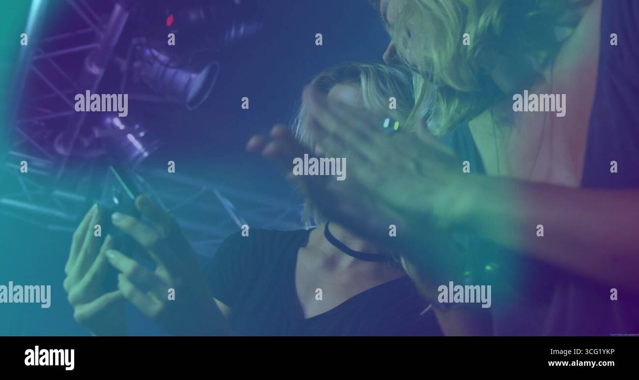 Scrolling smartphone by woman wearing choker and friend at club under truss haze spotlights Stock Photo