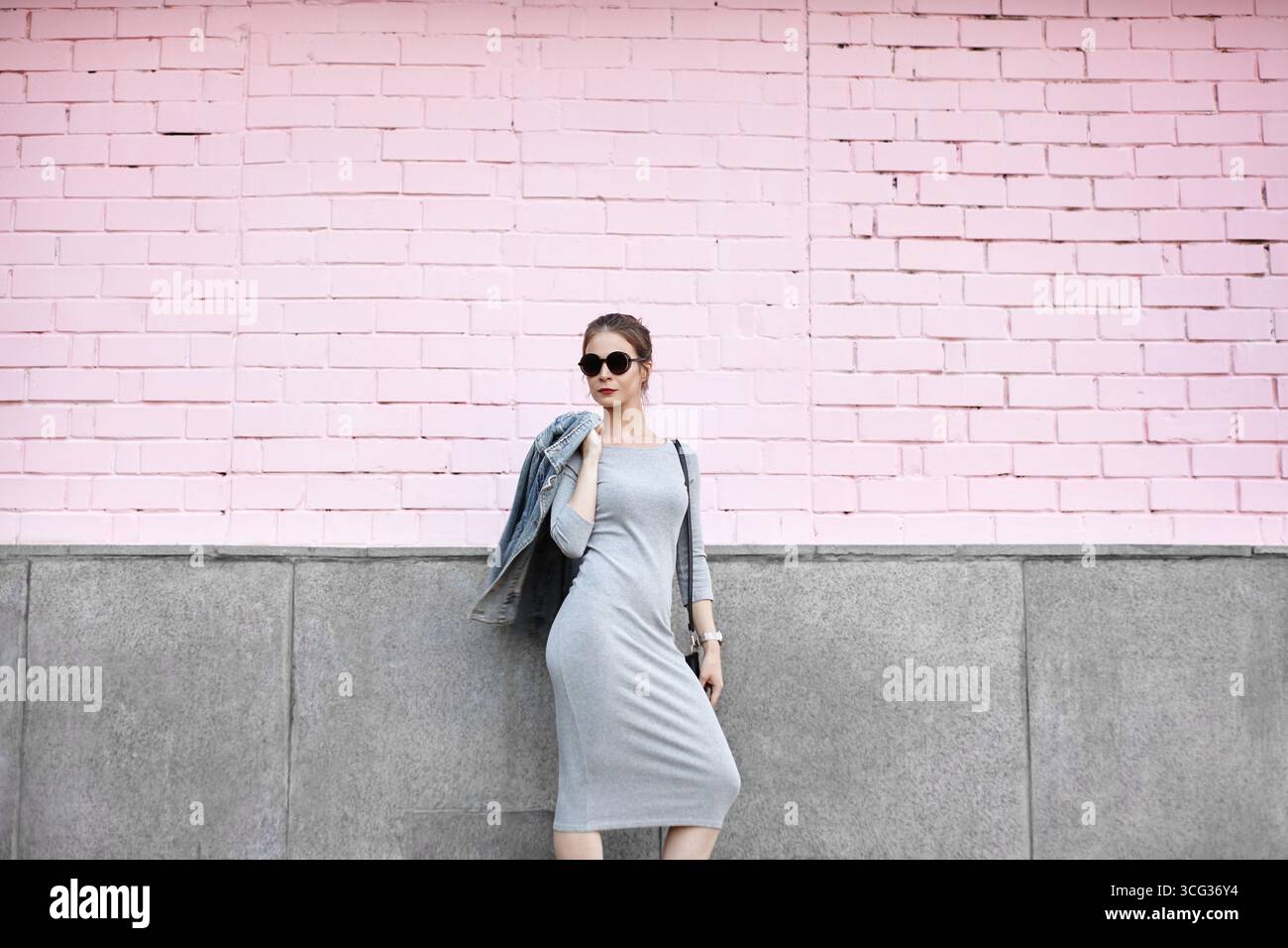 Stylish traveler woman in casual grey dress and sunglasses, leaning on pink wall under sunlight. Summer holidays lifestyle and outdoor adventure Stock Photo