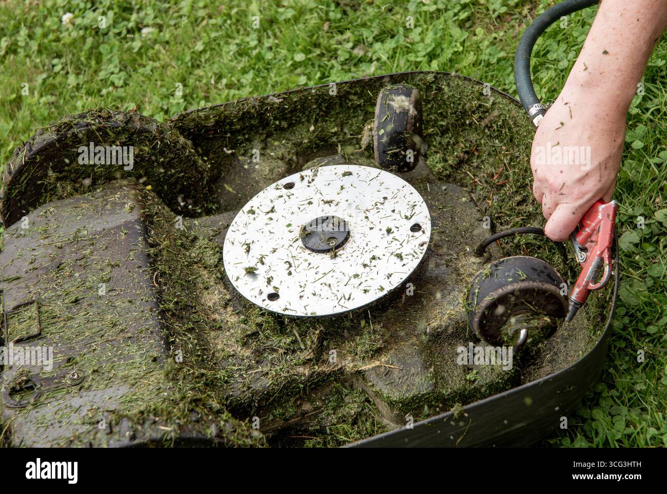 Close-up view of a man's hand blowing off a layer of wet grass stuck under an automated lawnmower using an air compressor — maintenance concept. Stock Photo