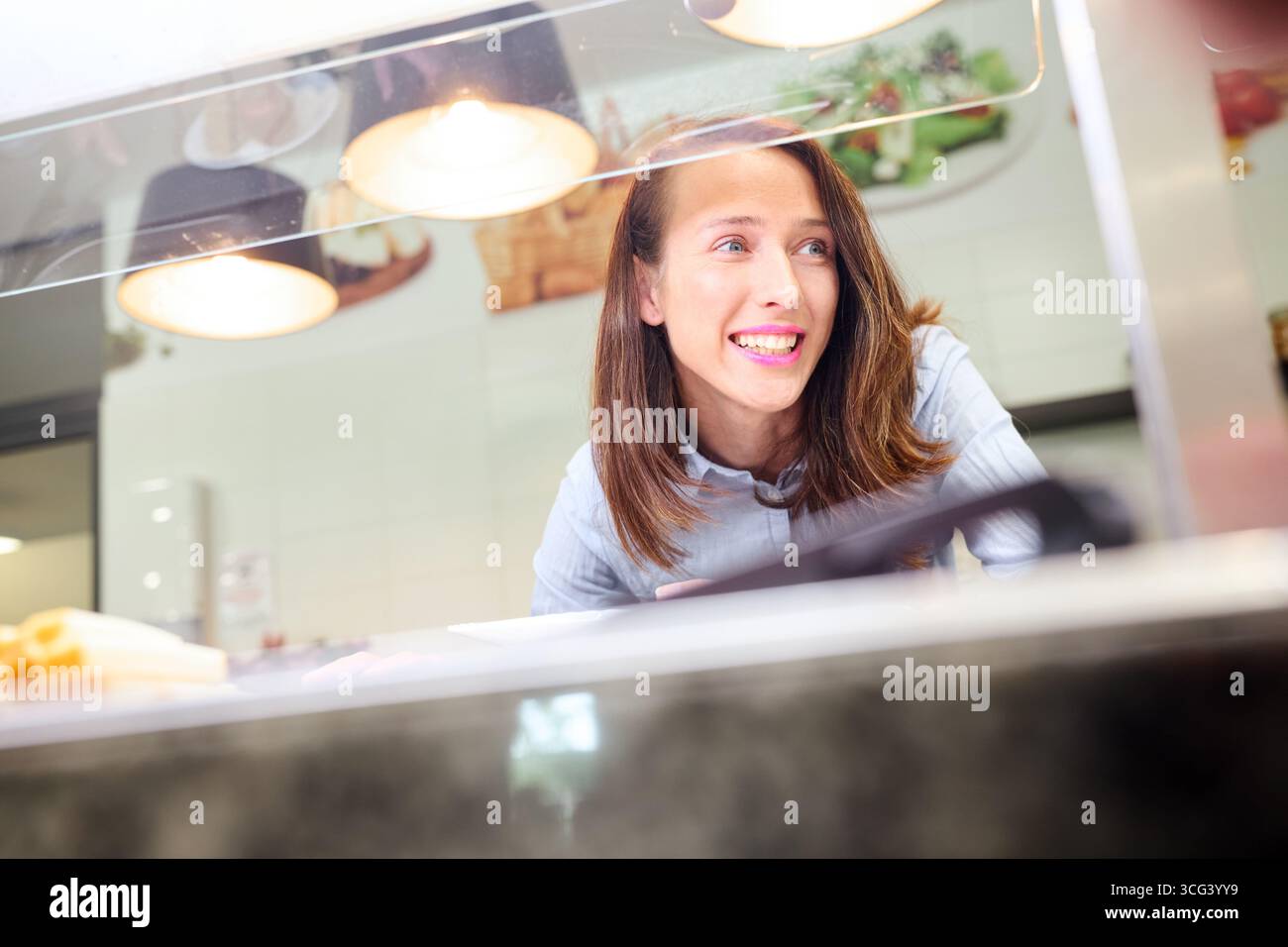 A cheerful woman standing behind a cafeteria counter in a clean and welcoming school setting, ready to serve meals, conveying a sense of hospitality a Stock Photo