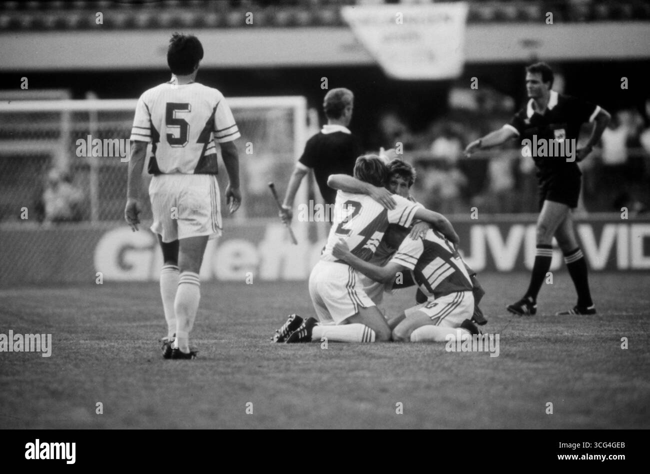 Drei Spieler der jugoslawischen Nationalmannschaft umarmen sich auf dem Rasen nach dem Einzug ins Viertelfinale der WM 1990. Faruk Hadzibegic schreitet auf die drei zu. Hinter ihnen geben sich Schieds- und Linienrichter die Hand. Stock Photo