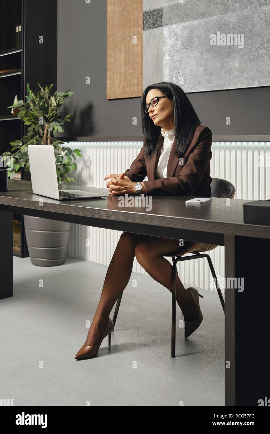 A woman dressed in business attire sits at an office desk in office and looking at the screen of laptop during a video call Stock Photo