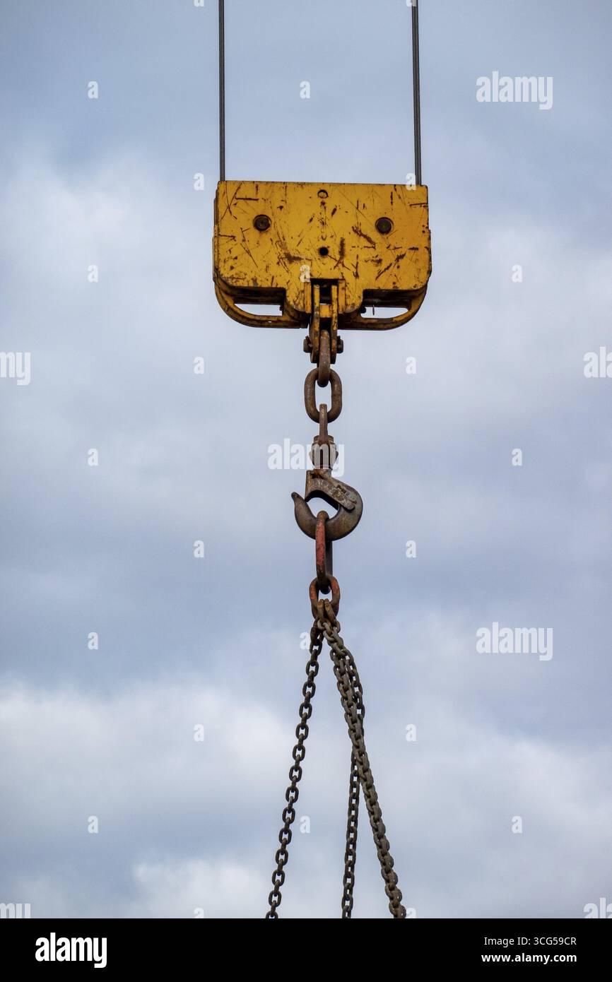 Rusty crane hook hanging from chains with cloudy sky background, representing industrial work and heavy lifting Stock Photo
