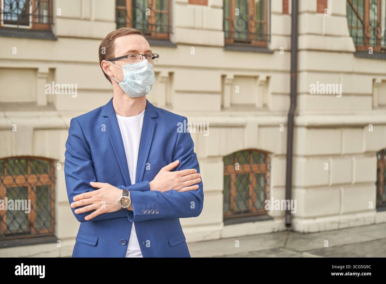 Young caucasian male wearing mask and blue jacket outdoors near building. Stock Photo