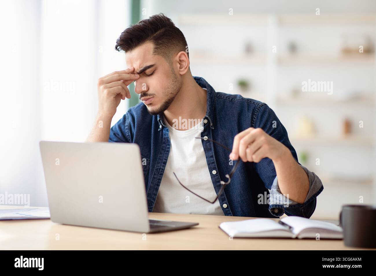 Exhausted arab guy sitting at workdesk, removing glasses Stock Photo