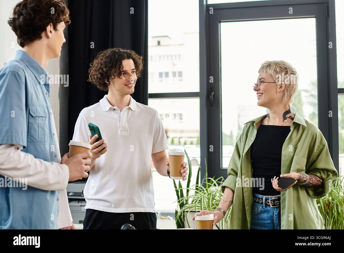 Three young professionals share coffee and ideas in a lively office while collaborating. Stock Photo