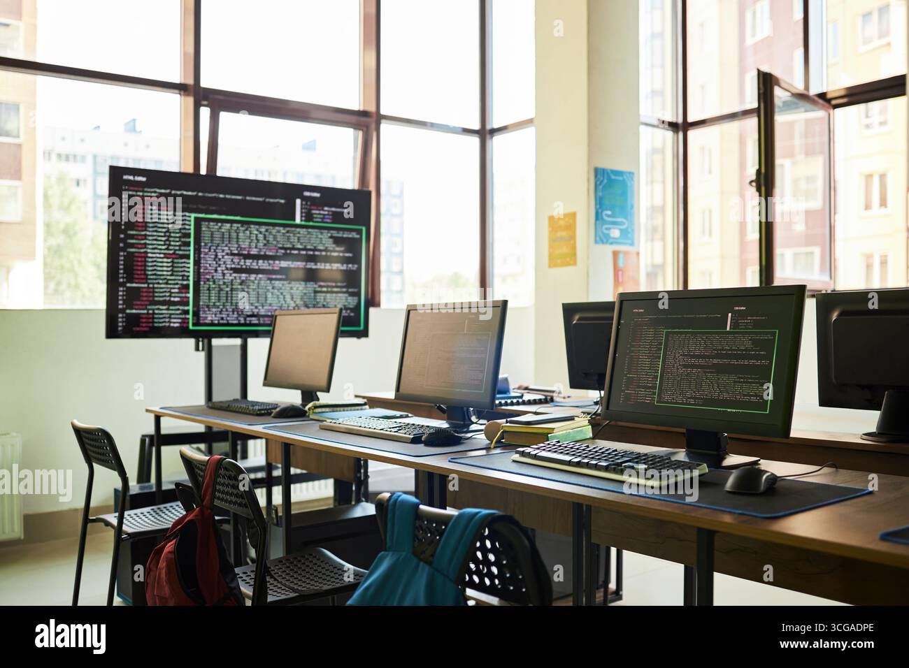 Empty classroom with multiple computer workstations displaying programming code on monitors, modern educational setting with large windows and backpacks on chairs Stock Photo
