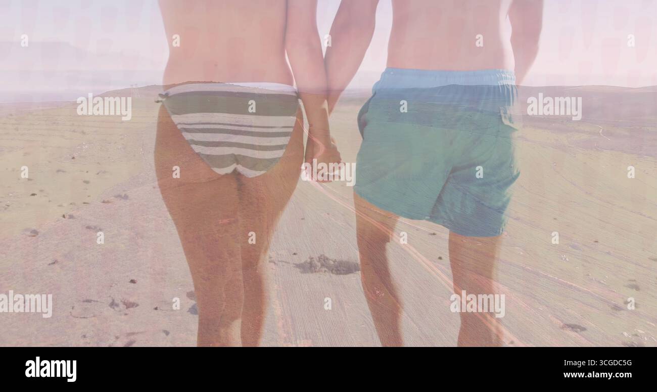Walking couple strolling hand in hand at sandy beach wearing striped bikini bottoms and swim shorts Stock Photo