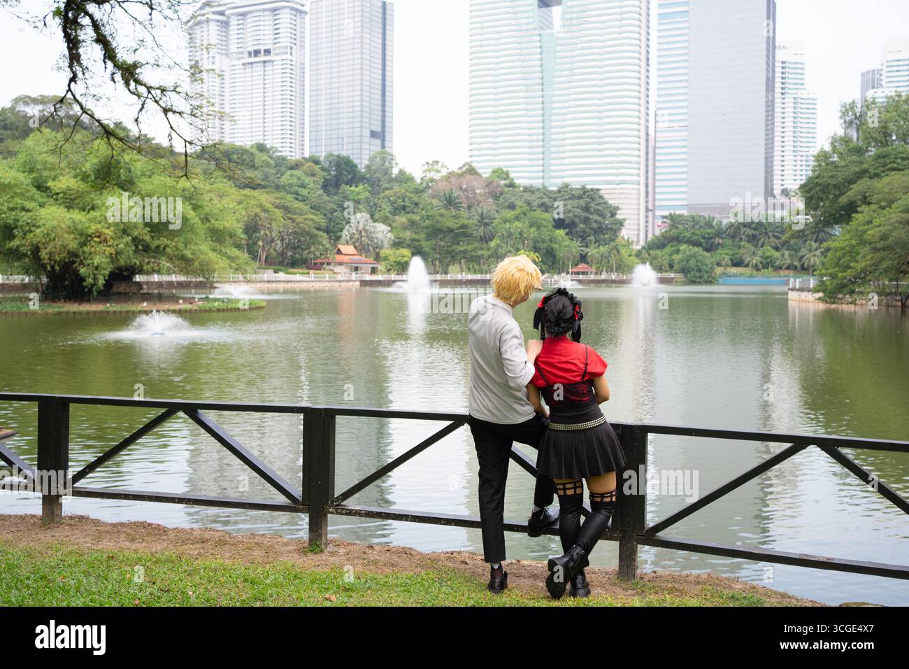 Two cosplayers admire a lake with fountains in a city park, surrounded by modern skyscrapers Stock Photo