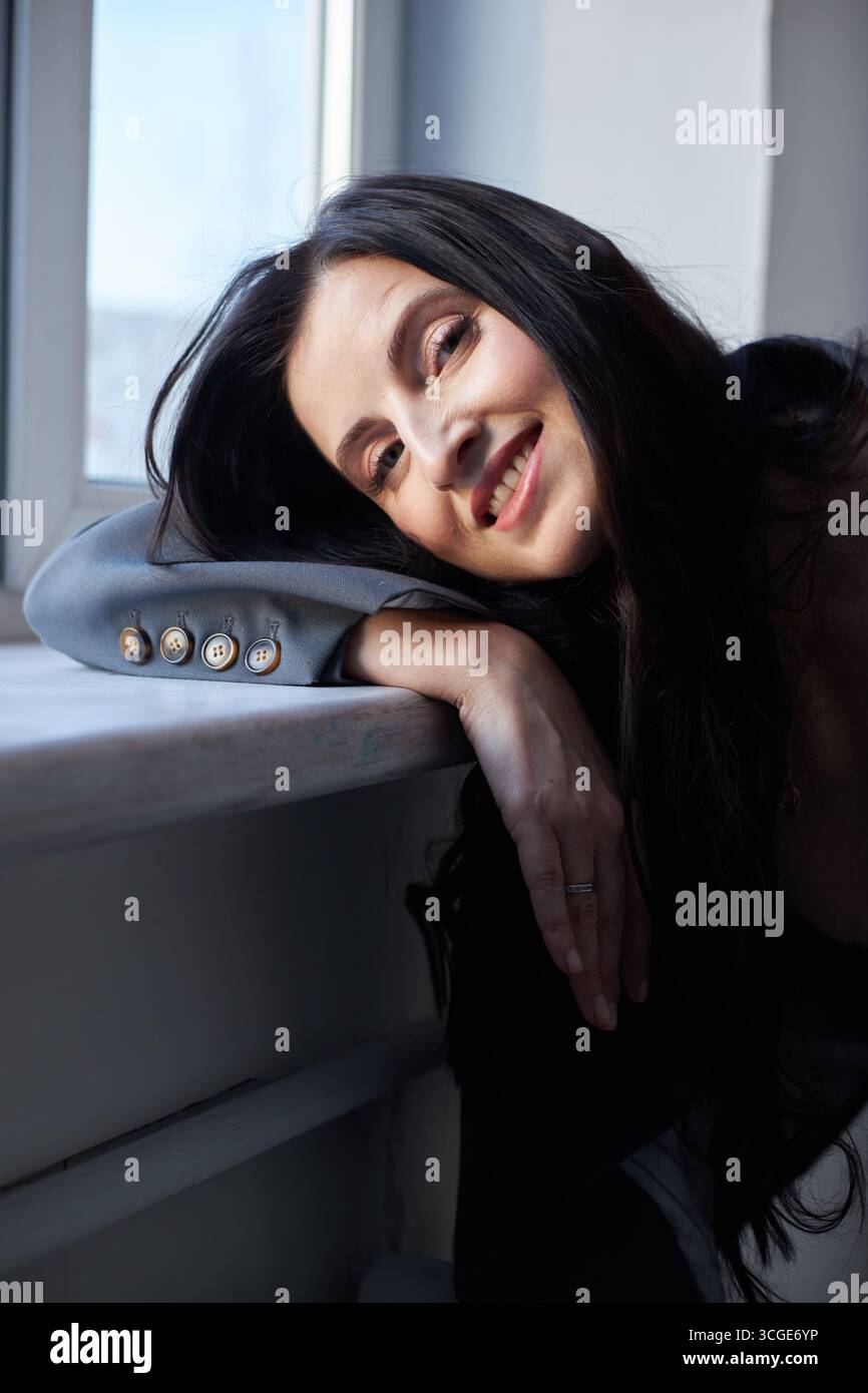 A young woman with long dark hair leans comfortably on a windowsill ...