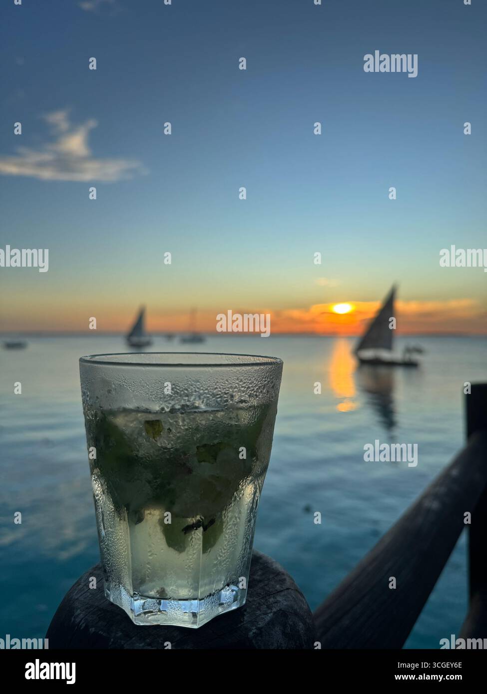 Refreshing cocktail glass with lime and mint overlooking ocean horizon and sailboats during sunset in Zanzibar, Tanzania, capturing holiday lifestyle vibes - Smartphone Captured Stock Image