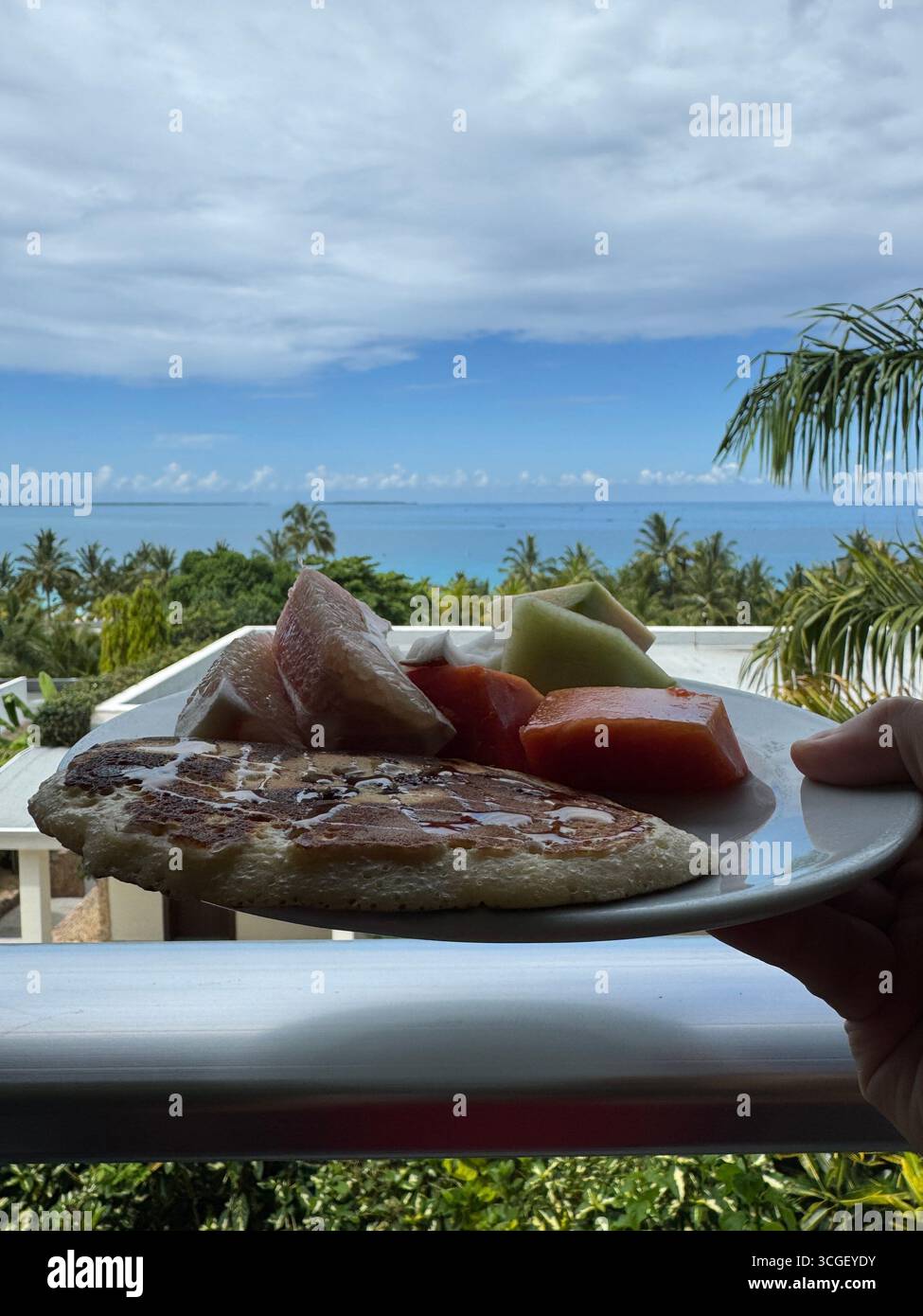 Fresh tropical breakfast plate of fruit and pancake overlooking lush greenery and ocean horizon in Zanzibar, Tanzania, capturing holiday resort lifestyle - Smartphone Captured Stock Image