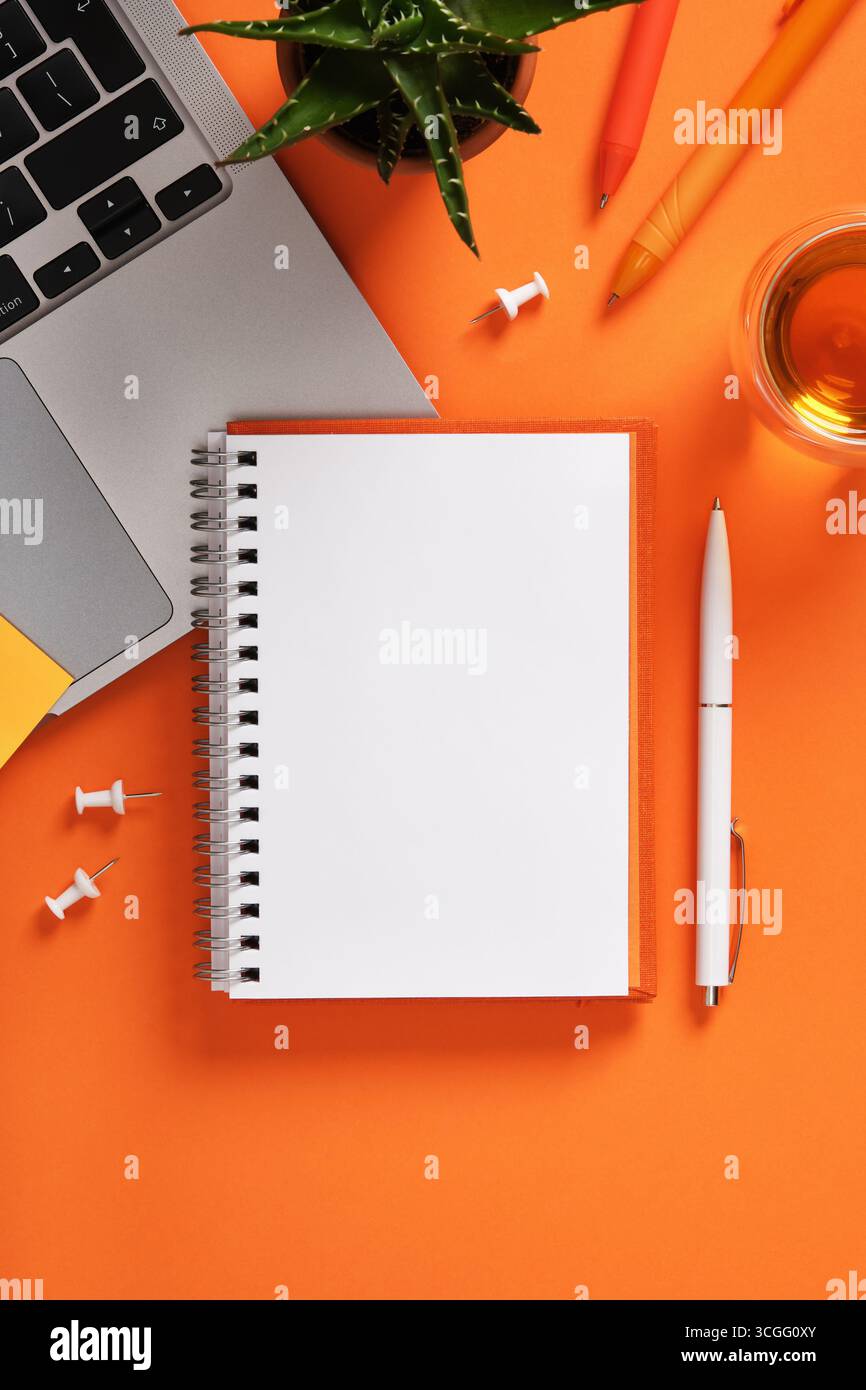 Blank notebook with laptop keyboard, pens, and cup of tea on orange desk. Workspace for study, planning, and creativity. Stock Photo