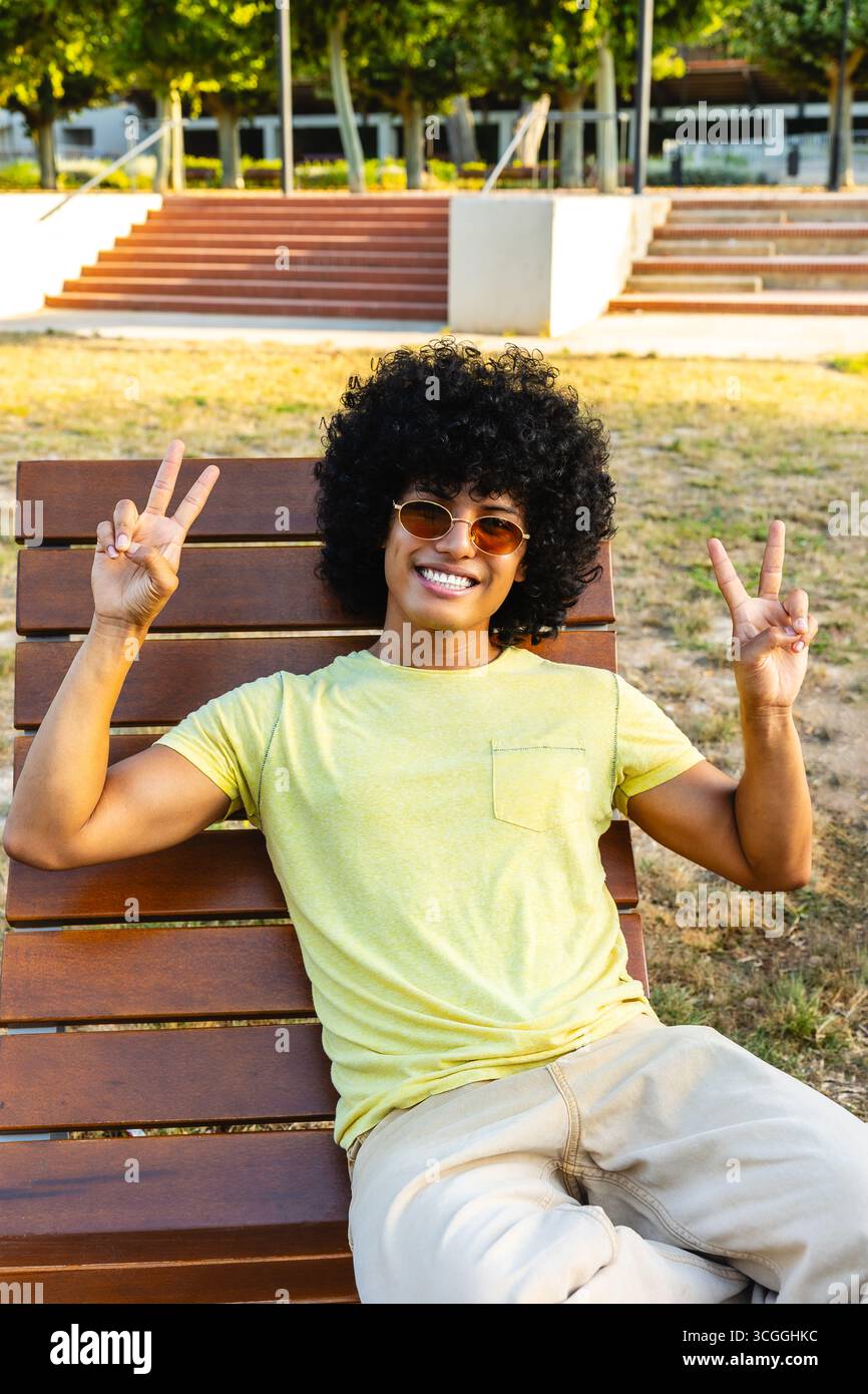 Young man showing peace sign while relaxing on bench in park Stock Photo